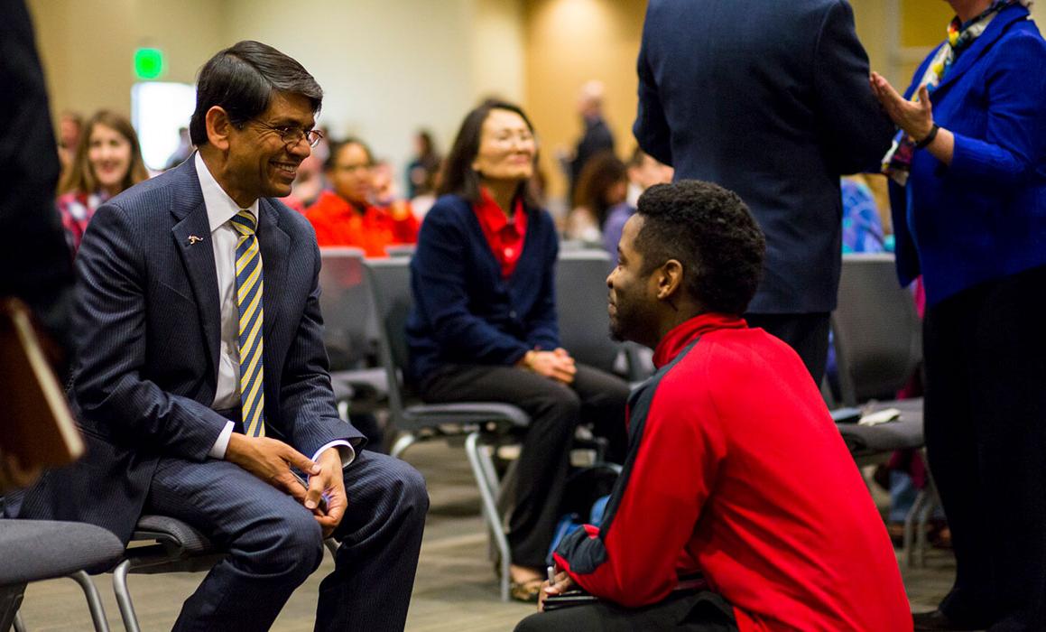 Student presenting as black male squats in front of seated and smiling Chancellor Agrawal