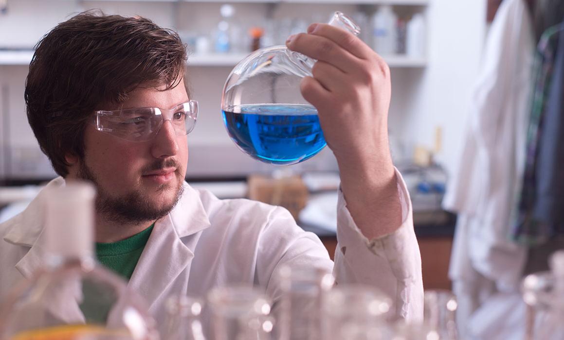 在实验室里，一名身着实验服、戴着安全眼镜的白人男性学生举起并看着装有蓝色液体的圆形小瓶