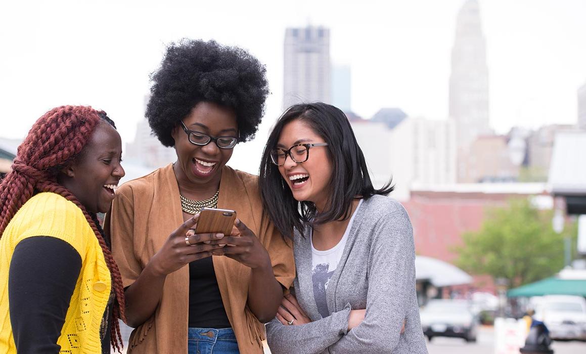 Three students presenting as female, two of whom present as Black, smile and laugh while looking at a phone together with KC skyline in background